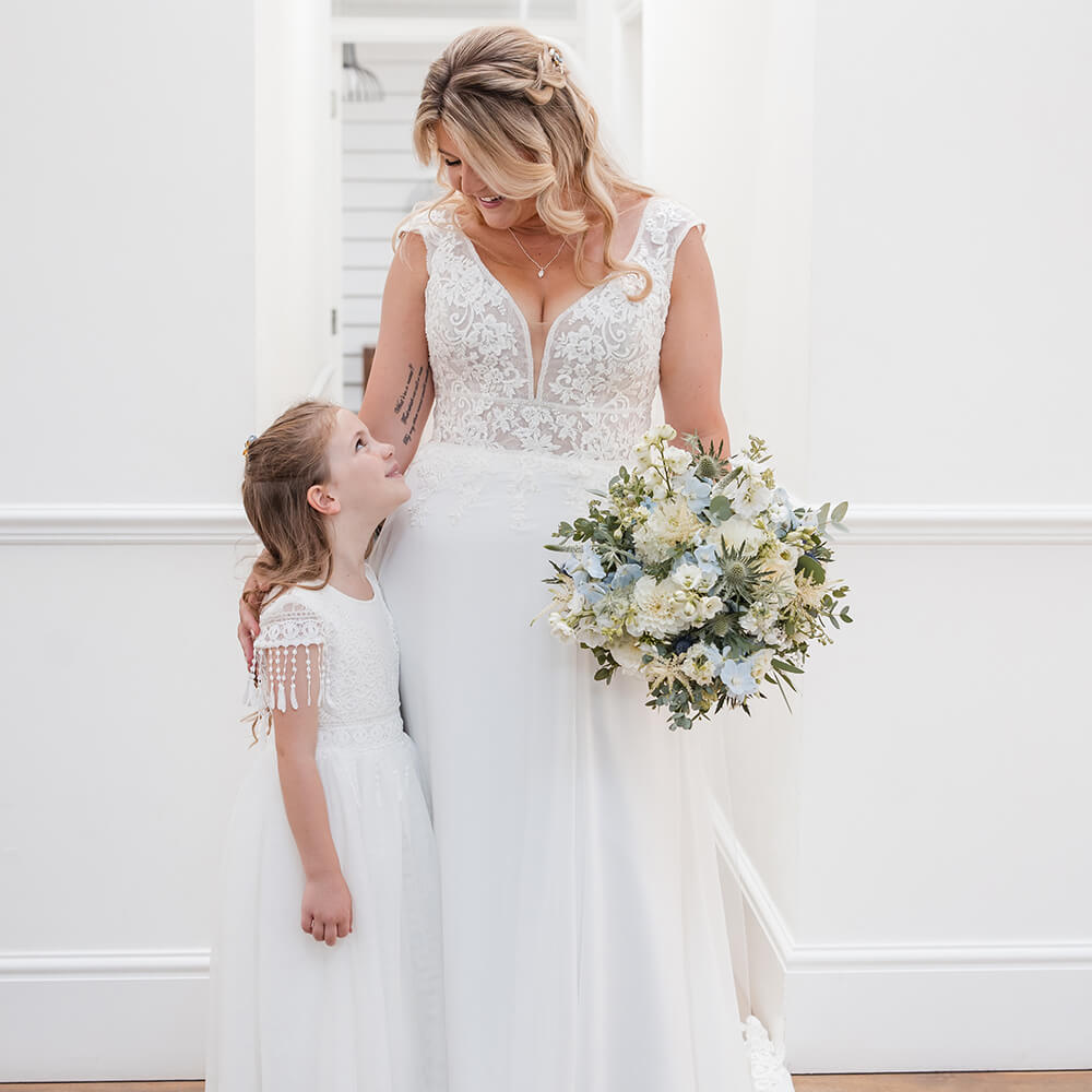 Woman in a wedding dress wearing wedding jewellery holding flowers and standing next to a young girl in a white dress